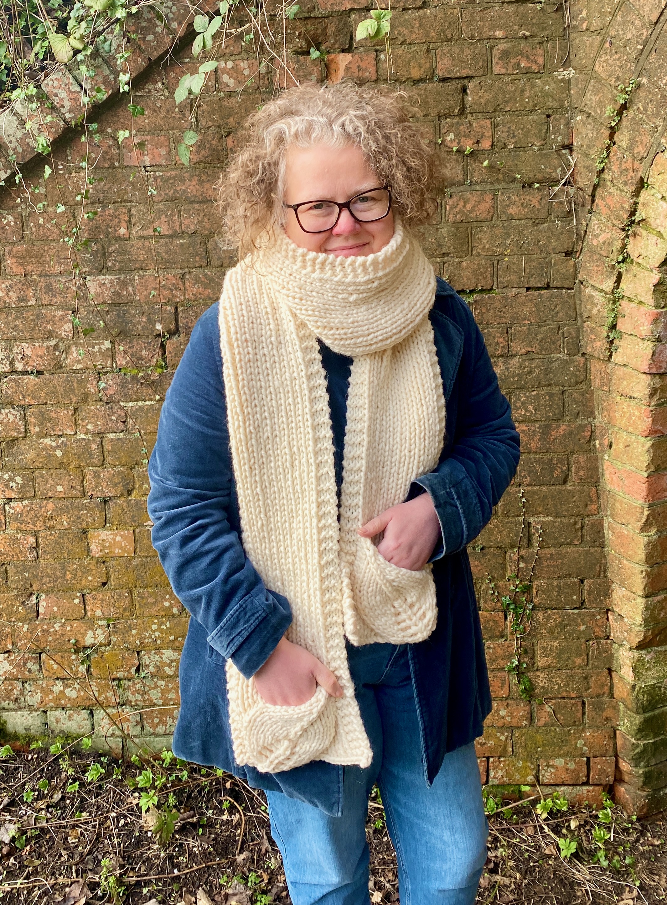 Hannah wearing a long wintery Cream scarf with pockets in front of an ivy covered wall
